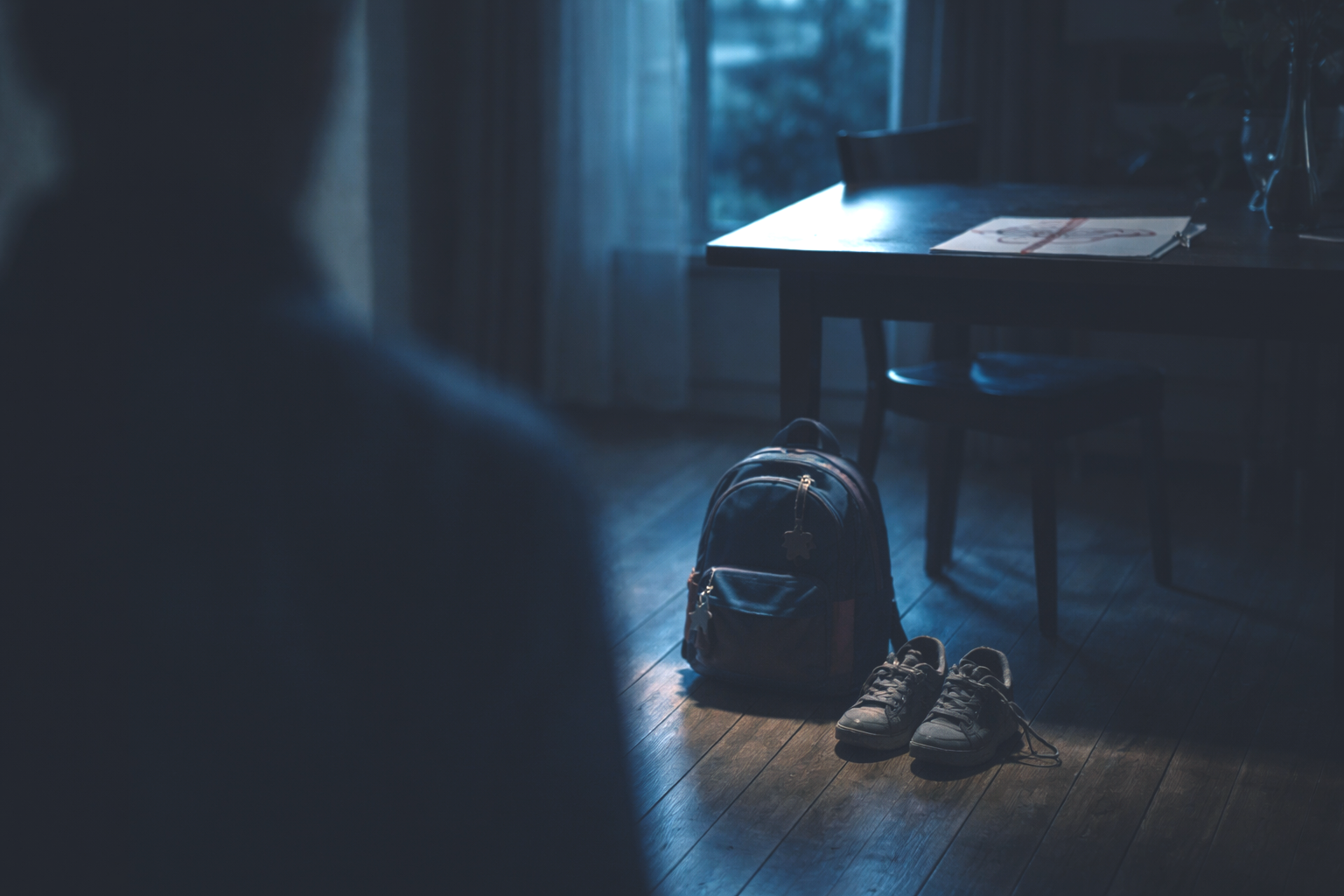 Interno domestico al crepuscolo con zaino e scarpe di un bambino sul pavimento, silhouette adulta sfocata, simbolo di pressione educativa e violenza psicologica